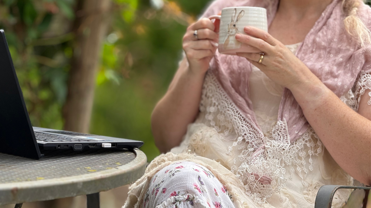 Person sitting outdoors with a laptop and mug, wearing a pink lace top.